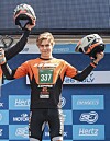 En ung motorsportutøver står på en pallen og holder en hjelm i hver hånd foran en sponsorvegg.