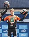 En ung motorsportutøver står på en pallen og holder en hjelm i hver hånd foran en sponsorvegg.