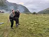 To personer bærer en sau over en grønn eng i fjellet med elv, bratte fjell og overskyet himmel.