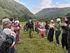 En gruppe mennesker står i en fjelldal og applauderer, omgitt av grønne fjellsider og skyet himmel.