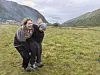 To personer står på en grønn eng i fjelldalen, med fjell og noen bygninger i bakgrunnen.
