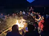 Folk sitter i ring rundt et kraftig bål om kvelden, med fjell og telt i bakgrunnen.