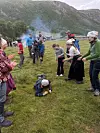 Gruppe mennesker står på en gresslette i fjellet med røyk i bakgrunnen; et barn ligger på bakken i forgrunnen.