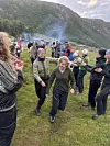 En gruppe mennesker står samlet på en gresslette i fjellet, mens røyk stiger opp fra et bål.