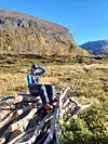 Person sitter på en stabel med ved i et åpent fjellområde med bratte fjellsider og blå himmel.