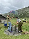 To personer står ved en stor metallkjele ved en seter med fjell og beitende kyr i bakgrunnen.