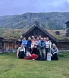 En gruppe mennesker står og sitter foran en gammel tømmerbygning med torvtak i et fjellområde.