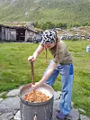 En person rører i en stor gryte med mat ved en seterbygning på en grønn fjelleng.