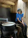 En person sitter ved en stor svart gryte og rører i væsken inne i et lite trehus.