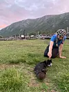En kvinne i blått sitter på gresset og peker mot en svart hund i et grønt landskap med fjell i bakgrunnen.