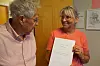 To eldre personer står innendørs, og en kvinne holder opp et dokument mens de smiler til hverandre.