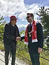 To menn håndhilser utendørs ved et rekkverk, med skyet himmel, fjell og vann bak.
