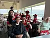 Flere eldre personer sitter i et lyst rom med store vinduer, noen i røde supporterhatter og drakter.