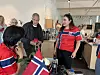 Flere personer står rundt et bord med norsk flagg, kaffeutstyr og kopper i et lyst rom.