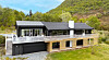 Moderne enebolig med mørkt tak, stor terrasse, garasje og grønn ås i bakgrunnen.