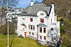 Hvit villa med røde vindusrammer i en grønn skråning, med vann og andre bygninger i bakgrunnen.