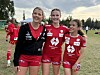 Tre unge fotballspillere i rødt og svart står smilende på en gressbane.