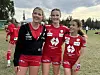 Tre unge fotballspillere i rødt og svart står smilende på en gressbane.