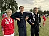 Tre unge fotballspillere står på en gressbane med mål og flere spillere i bakgrunnen.