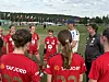 Jentefotballag i røde drakter står samlet i en sirkel på en gressbane med trenere rundt.