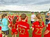 Flere jenter i røde og turkise fotballdrakter står samlet på en gressbane med trenere i bakgrunnen.