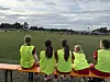 Fire jenter i gule vester sitter på en benk og ser ut over en fotballbane.