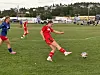 To fotballspillere i røde drakter og én i blå drakt på en gressbane med ballen i spill.