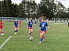 Ungdomsspillere i blå og røde fotballdrakter på en gressbane med mål og trær i bakgrunnen.