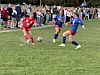 Tre fotballspillere i rødt og blått kjemper om ballen på en gressbane med publikum i bakgrunnen.