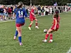 Tre jenter i røde og blå fotballdrakter spiller på en gressbane med publikum langs sidelinjen.