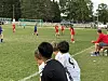 Ungdomsspillere i røde og blå drakter på en fotballbane med mål i bakgrunnen og folk langs sidelinjen.