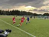 To spillere i røde drakter og en i blått løper etter ballen på en fotballbane.