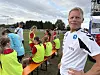 En trener står foran en benk med unge fotballspillere i gule vester på en grusbane.