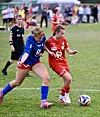 To fotballspillere i blått og rødt kjemper om ballen på en gressbane, med publikum i bakgrunnen.