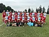 En stor gruppe jenter i røde fotballdrakter står og sitter samlet på gresset før kamp.