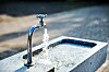 Metallkran i en steinfontene med vann som spruter ut i dagslys.
