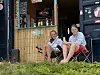 To ungdommer sitter på stoler foran en strandbod med bambusvegg og utstyr.