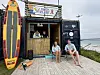 En liten strandkiosk med surfebrett, utstyr og to personer som sitter på en treplattform ved sjøen.