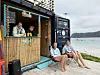 To personer sitter på benker ved en strandbod, med sjø og flere badende i bakgrunnen.