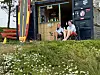 To ungdommer sitter foran en liten bod med surfebrett, utstyr og en strandkant med gress og blomster.