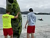 To personer bærer et grønt vannsportbrett på en strand med sjø og småbåter i bakgrunnen.