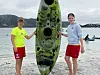 To ungdommer står ved en grønn kajakk som er reist loddrett på en strand, med vann og fjell i bakgrunnen.