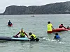 Flere barn og ungdom er i vannet ved en strand, med kajakk, brett og røde oppblåsbare flåter.