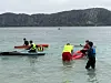 Flere personer står i vannet ved kajakker og utstyr nær en kyst med fjell i bakgrunnen.