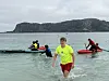 Flere ungdommer i neonfargede vester padler og står i vannet ved en strand.
