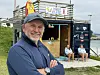 En mann står foran en liten strandkiosk med to personer som sitter på benker ved siden av.
