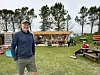 En mann står på gresset foran en rekke boder ved stranden, med trær, benker og folk i bakgrunnen.