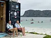 To personer sitter i campingstoler ved en strand med fjell, hav og flere padlere i bakgrunnen.