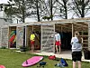 Flere små boder med utstyr ved en strand, med ungdommer og en person med kamera foran.