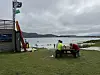 En strand med vannsportbod, to personer ved et bord og rolig sjø under overskyet himmel.